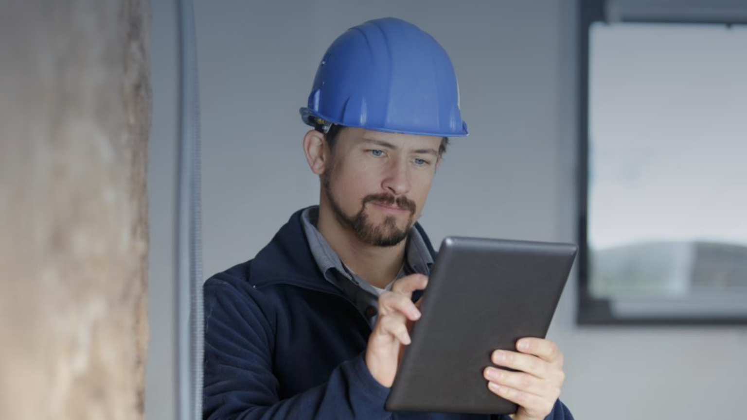 A man wearing a hard hat is holding a tablet computer, focused on his work in a construction or industrial setting.