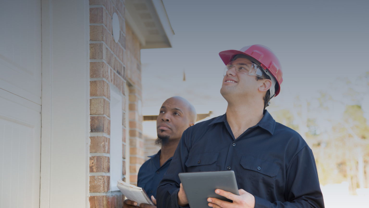 Two men in hard hats are examining a tablet, discussing plans or data on a construction site.