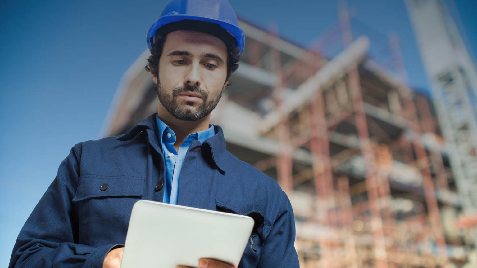 A man wearing a blue hard hat is holding a tablet, focused on his work in a construction or industrial setting.