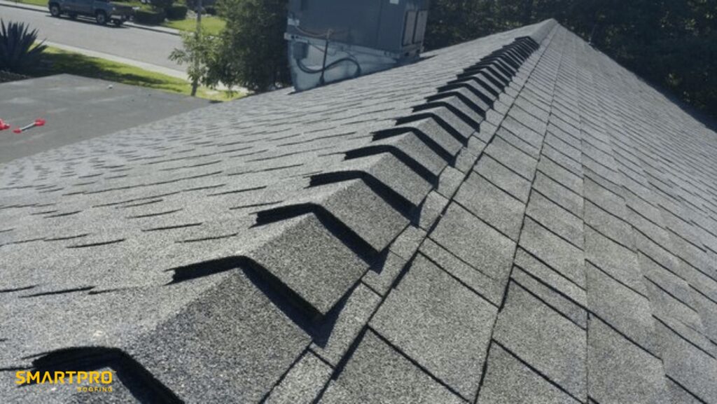Shingle Roof Lifespan Explained: How Long Do Shingles Last? (15, 20, and 30-Year Options) 11 Close-up view of a roof with dark asphalt shingles and ridge vent, taken from the peak looking down toward the street. "SmartPro Roofing" logo is visible in the bottom left corner.