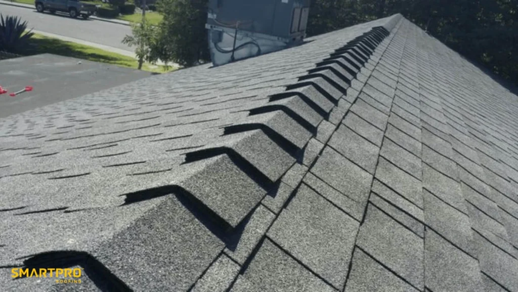 Close-up view of a roof with dark asphalt shingles and ridge vent, taken from the peak looking down toward the street. "SmartPro Roofing" logo is visible in the bottom left corner.