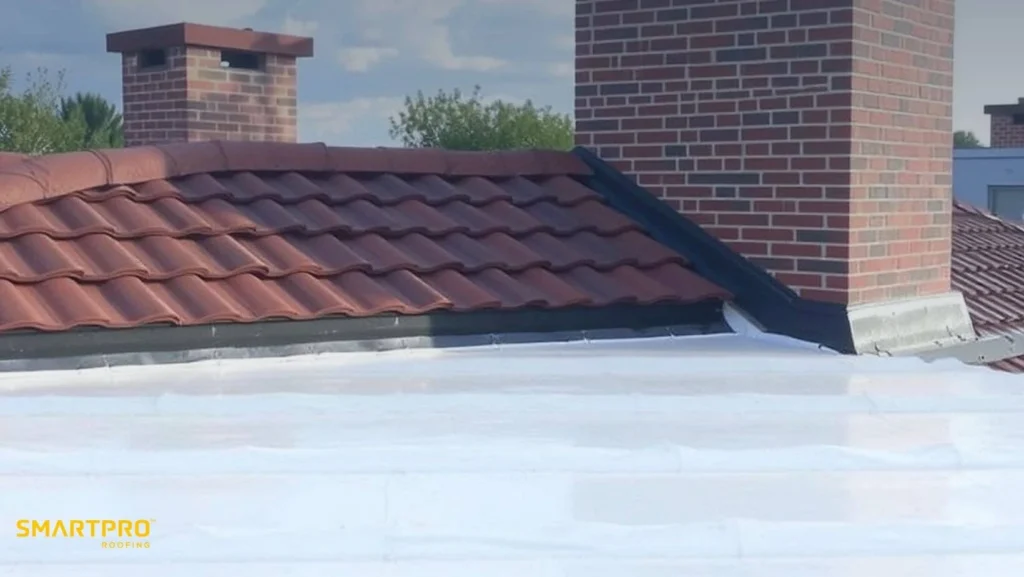 A rooftop scene featuring red clay tiles next to a brick chimney. The foreground shows a white flat roof. Logo text reads "SmartPro Roofing." Sky is partly cloudy.