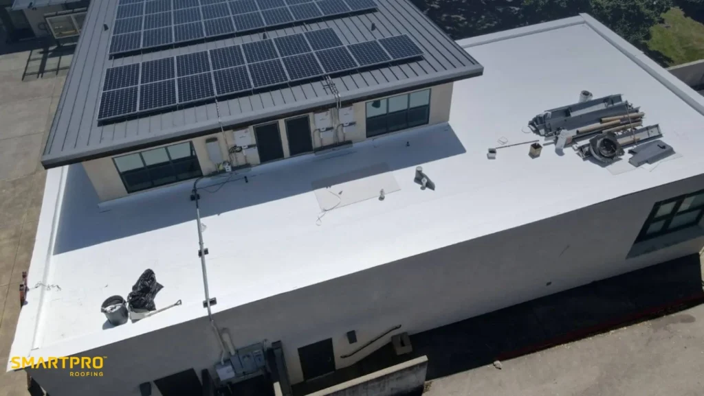 Aerial view of a building with a clean white roof, featuring solar panels on a section. Equipment and tools are scattered, indicating ongoing work. Bright and orderly.