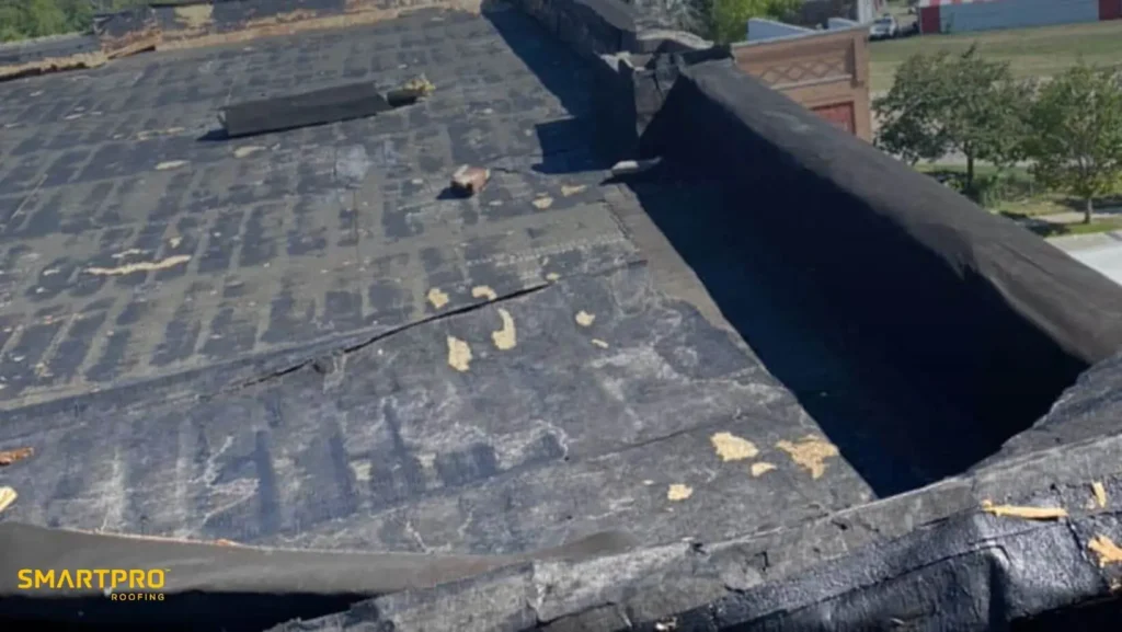 Flat Roof Leaks in Florida: Common Issues and How to Prevent Them 12 A flat rooftop with worn, black tar paper showing signs of weathering and damage. Sparse patches of trees and buildings are visible in the background.