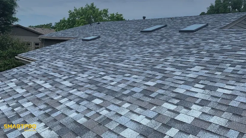 Alt text: "Roof with new gray asphalt shingles, featuring three skylights. Trees and parts of neighboring houses are visible under a cloudy sky."