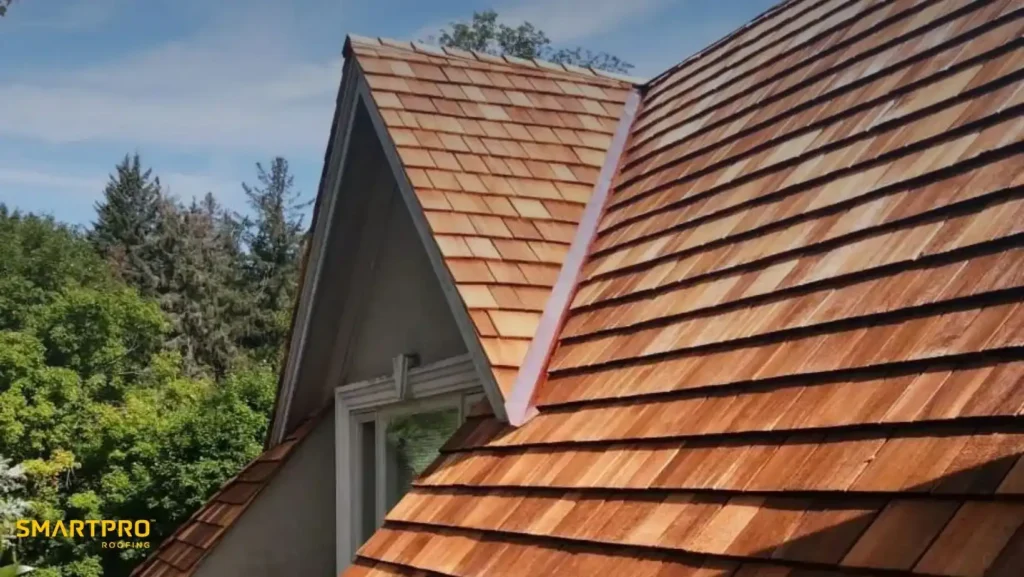 A sloping roof with new wooden shingles under a clear blue sky. A small window is visible, surrounded by lush green trees. Logo: SmartPro Roofing.