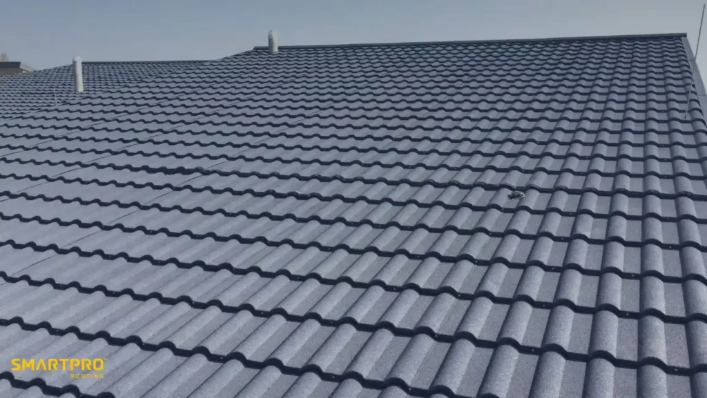 Steel Shingles: Durable, Stylish, and Long-Lasting Roofing Solutions 11 A rooftop with blue-grey, wavy-patterned tiles under a clear sky. Clean, orderly design with industrial chimneys visible. Logos in the bottom left.