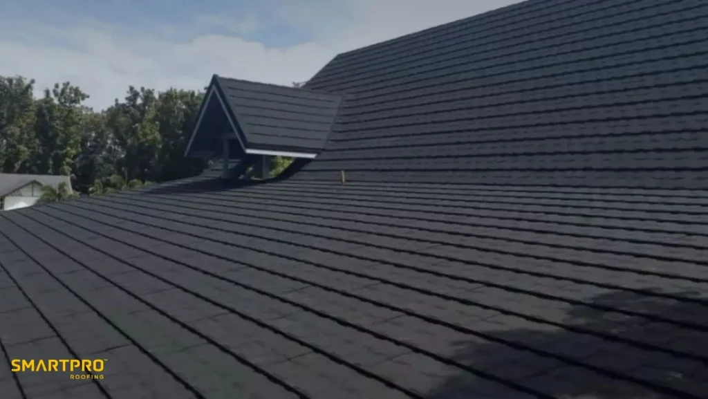 A sleek, dark metal roof with clean lines and a small dormer window reflects sunlight. Green trees line the background, under a clear sky. Logo reads "SmartPro Roofing."