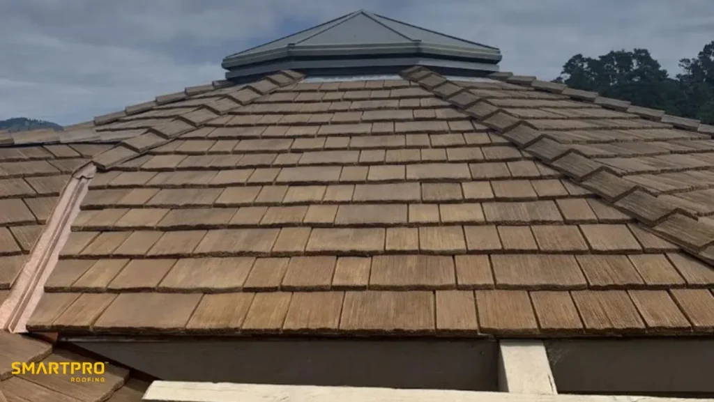 A close-up view of a wooden shingle roof with a light brown hue under a cloudy sky. Trees are visible in the background. The mood is calm and rustic.