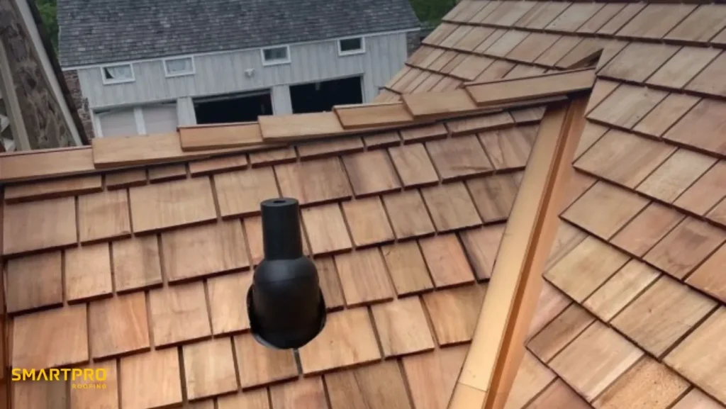 A wooden shingle roof with a neat, uniform pattern, featuring a black vent pipe near the center. The background shows a neighboring, similarly styled building.