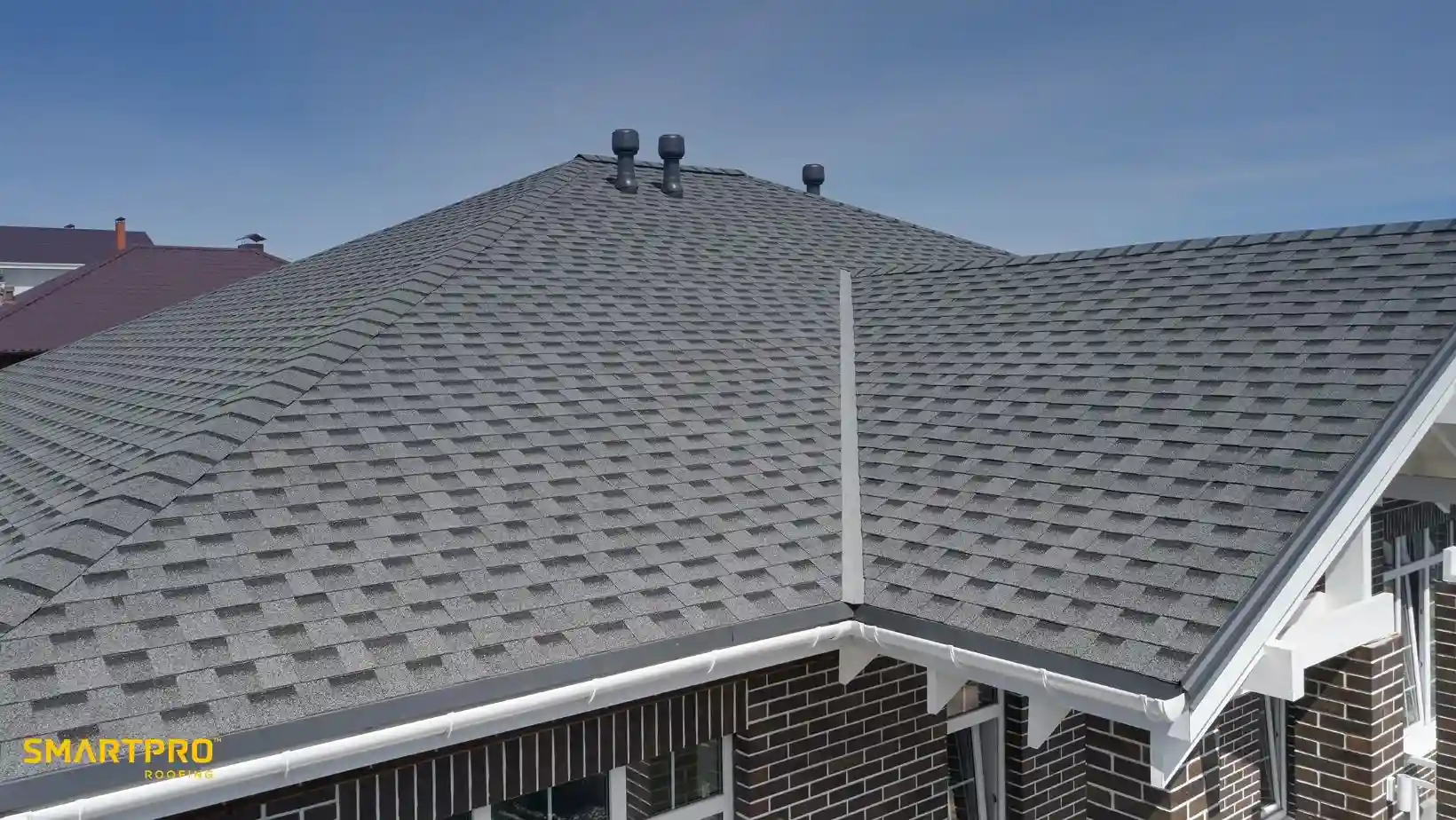 A modern house with a gray shingle roof, featuring multiple vents. The structure is made of dark bricks with white trim, under a clear blue sky.