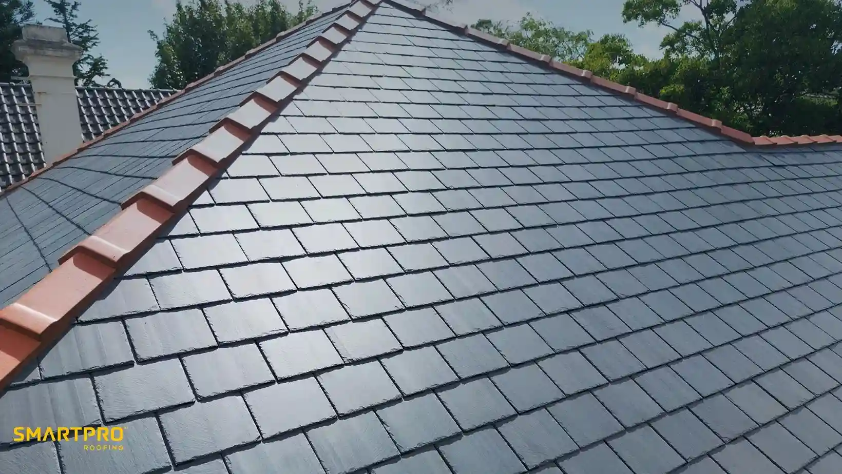 A sleek, newly-tiled gray slate roof with red ridge tiles under a clear blue sky. Lush green trees frame the background, evoking a sense of freshness.