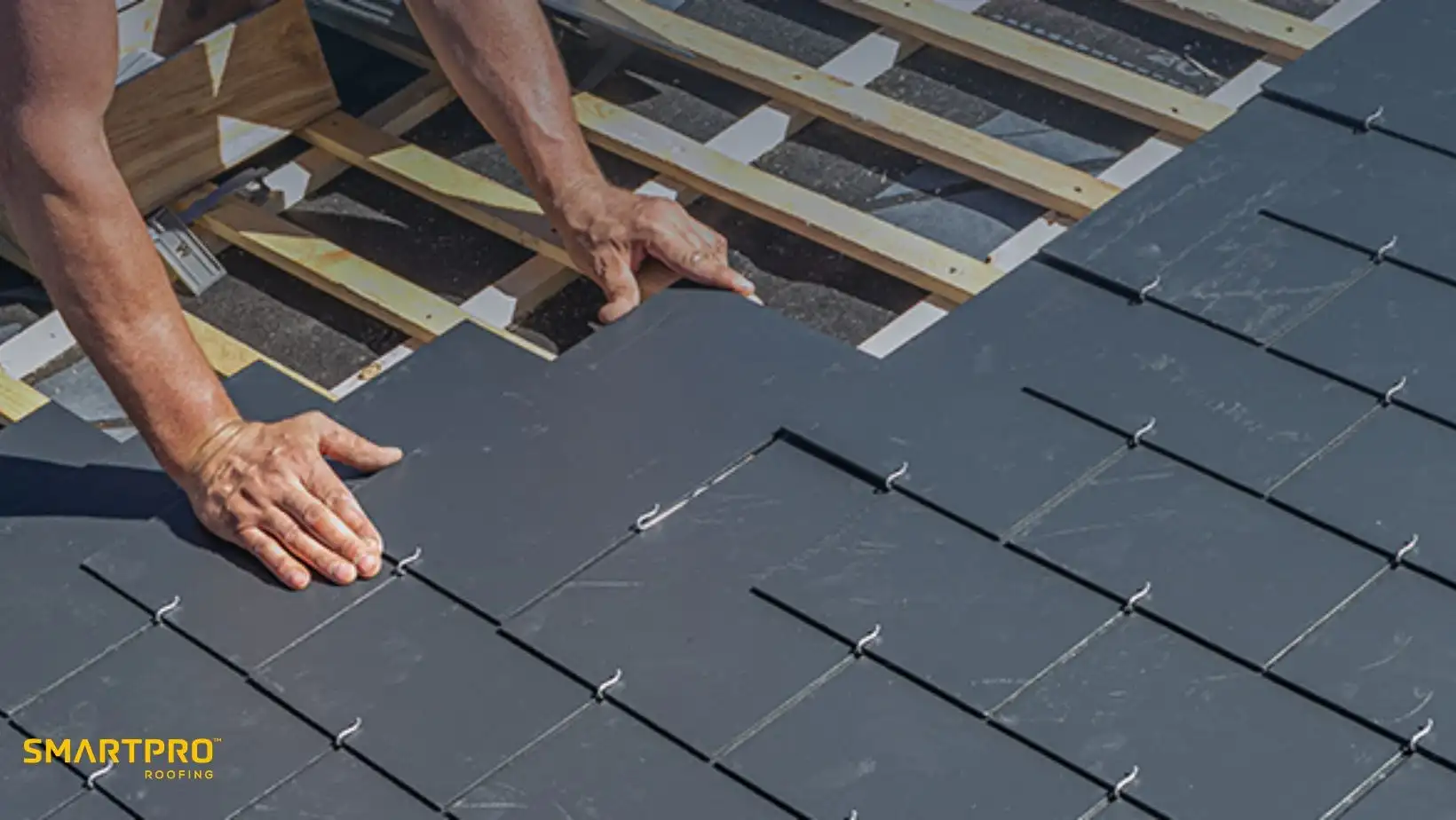 A person is installing dark slate roof tiles on a wooden framework, showcasing precise craftsmanship. The image conveys a sense of skilled labor.
