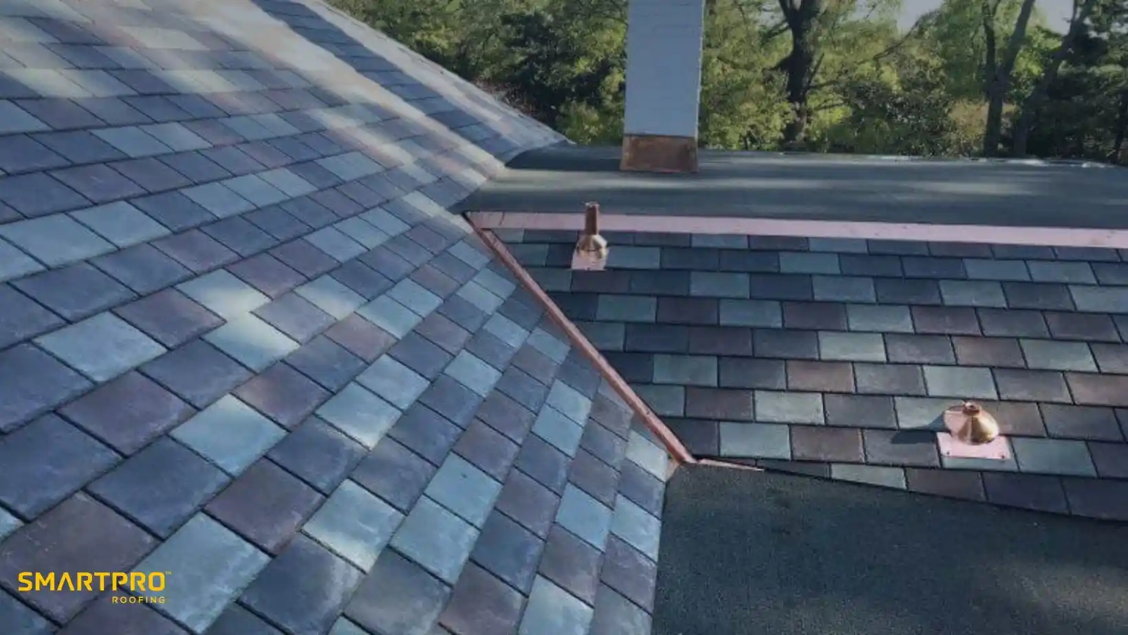 A rooftop with blue and gray slate tiles, showing clean lines and two small vents. Trees in the background suggest a serene, natural setting.