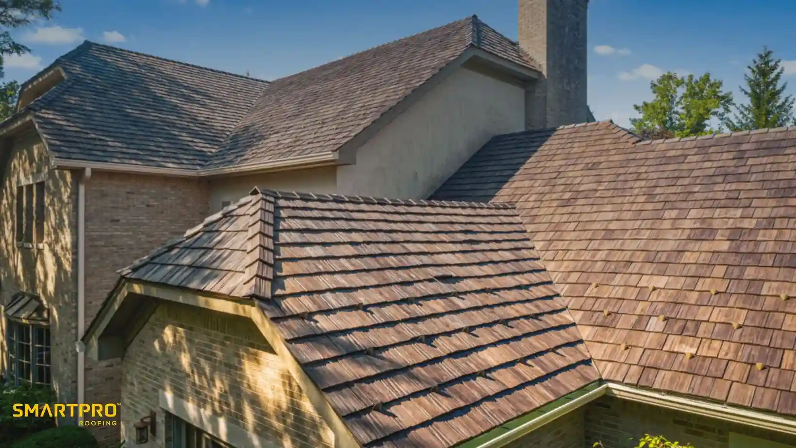 A large house with a multi-layered, brown shingle roof against a clear sky. The light stone walls contrast with green trim. A tree in the background suggests a suburban setting. The tone is serene and the roofing appears high-quality.