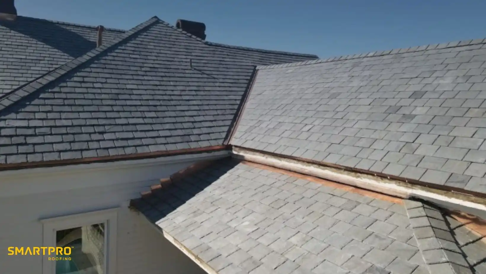 A close-up of a slate-tiled roof on a sunny day, showing clean, neatly arranged gray tiles. The roof edges have copper flashing. SmartPro Roofing logo in the corner.