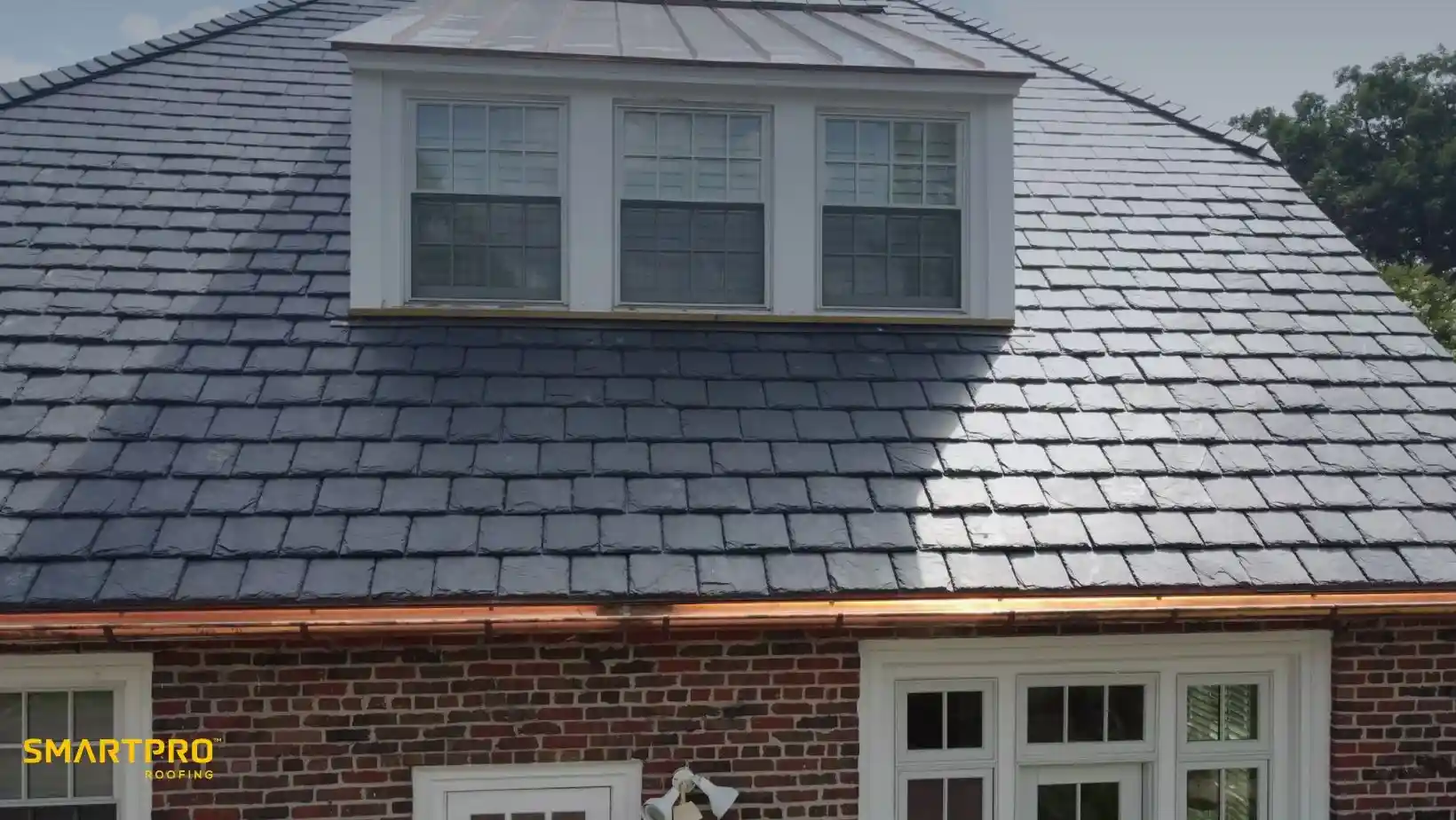 A brick house features a shiny slate roof with a central dormer window, exuding a modern and clean appearance. "Smart Pro Roofing" is in the corner.