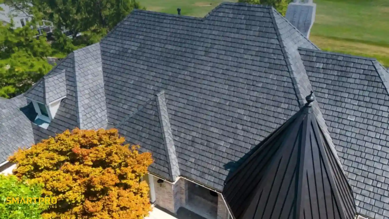 Aerial view of a house with dark gray, shingled, multi-angled roof, surrounded by trees. The roof conveys a modern and elegant design.