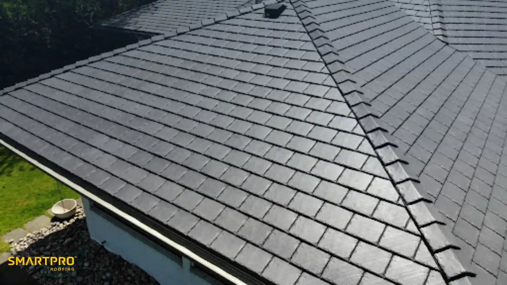 A sleek, modern black shingle roof with neat overlapping tiles in bright sunlight, bordered by green grass and a white gutter. SmartPro Roofing logo present.