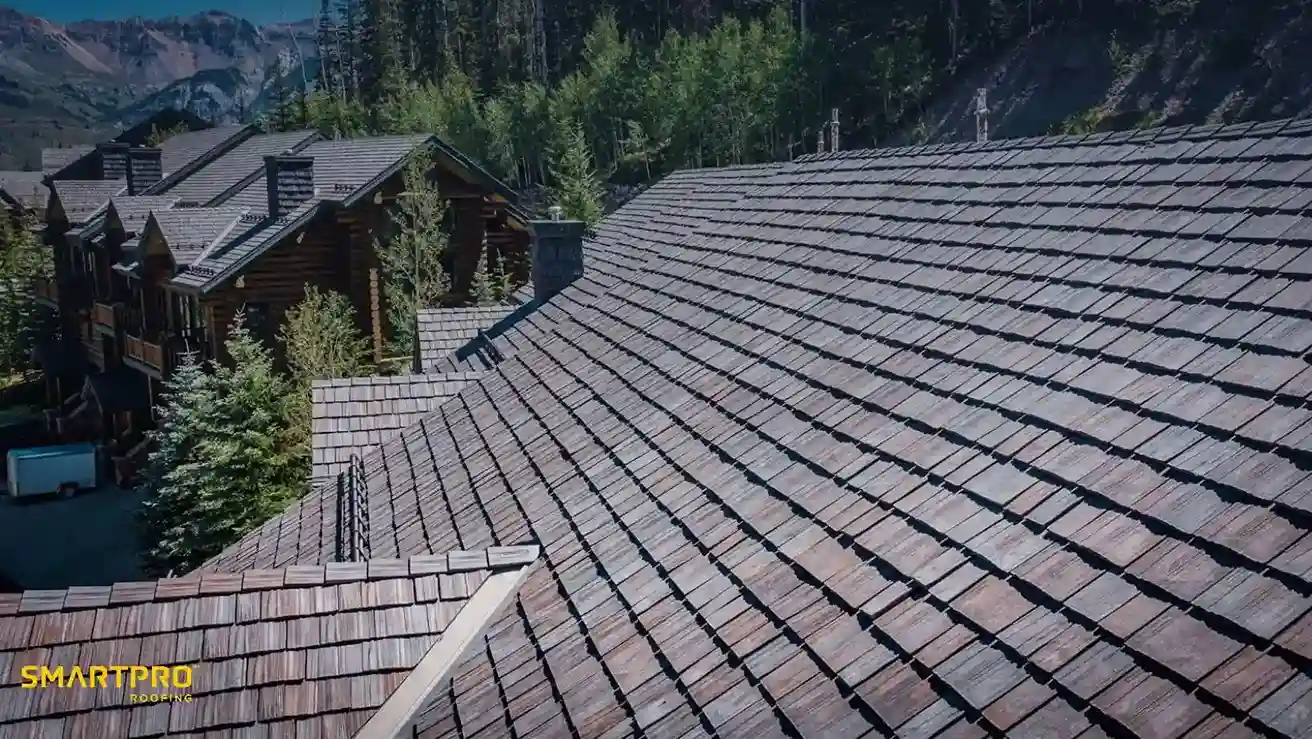 Aerial view of a large, sloped shingle roof against a backdrop of log cabins and pine trees. The setting is serene, nestled in a mountain landscape.