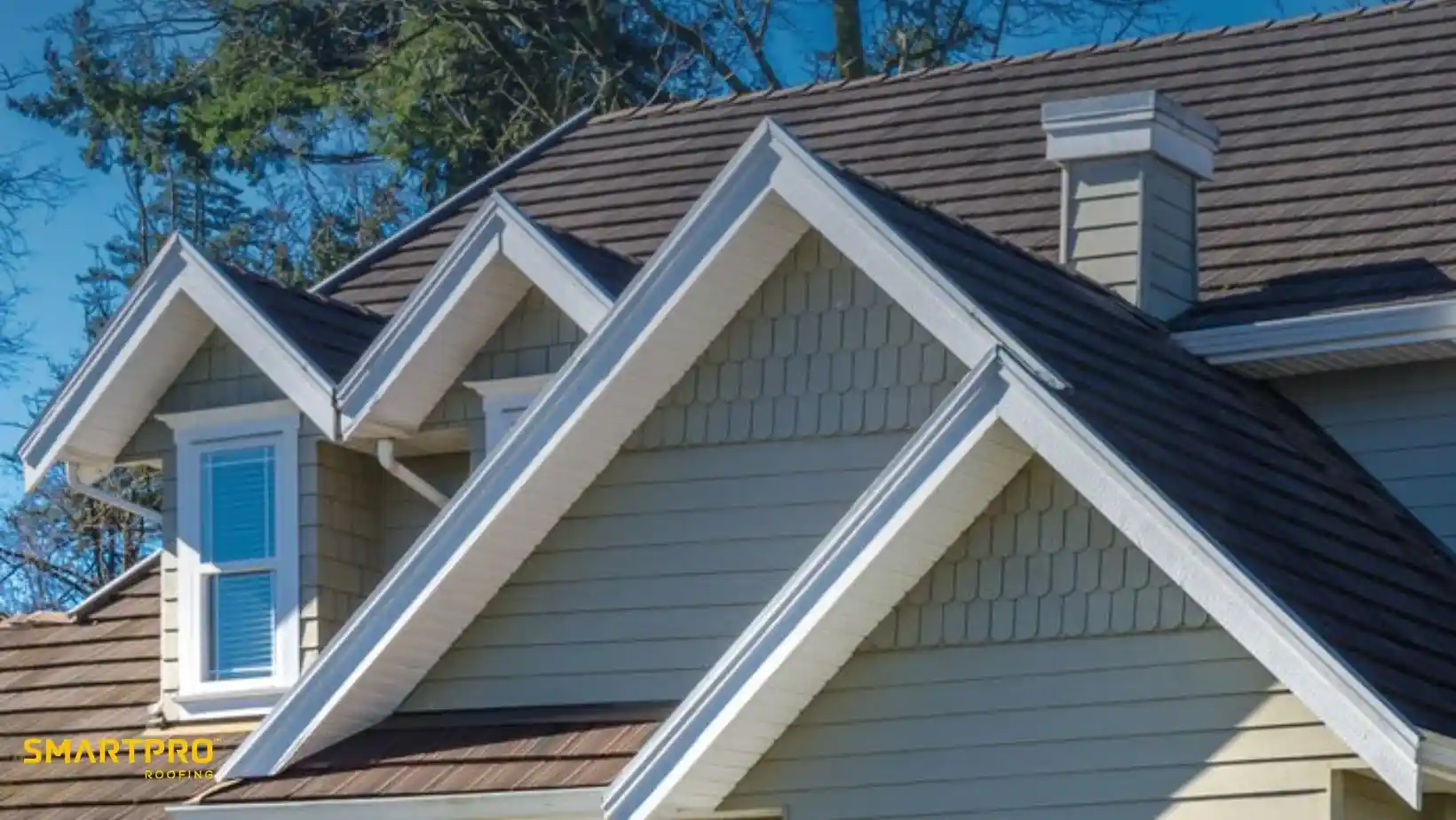 Close-up view of a house with beige siding, featuring multiple gabled roofs and white trim. A bright sky and trees form the backdrop.