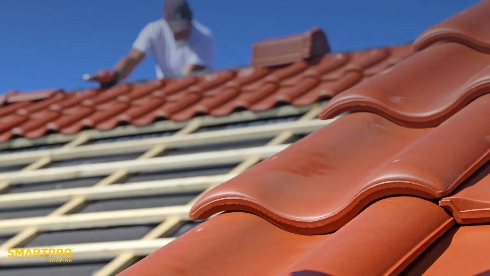 A worker installs red terracotta tiles on a roof under clear blue skies. Focus on the neatly aligned tiles, conveying precision and craftsmanship.