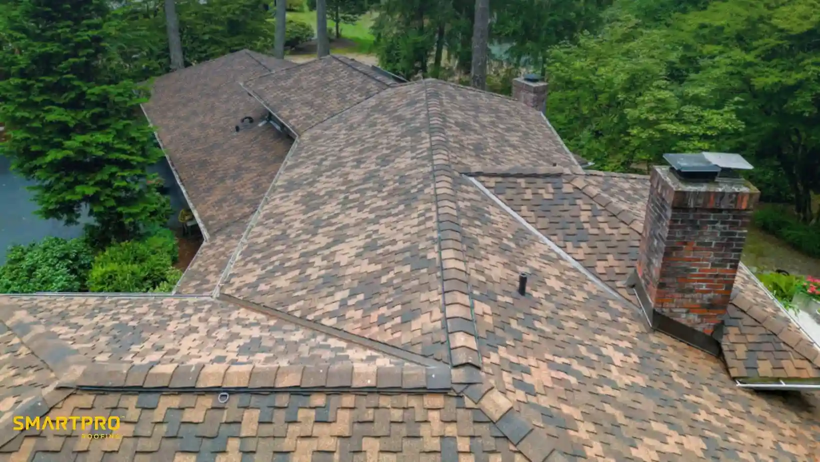 Aerial view of a large house with a new, brown shingle roof surrounded by lush green trees. A brick chimney is prominently visible, adding texture.