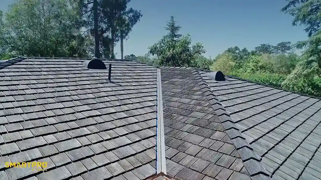 Aerial view of a wooden shingle roof with two vents, surrounded by lush green trees under a clear blue sky, conveying a serene, rural setting.