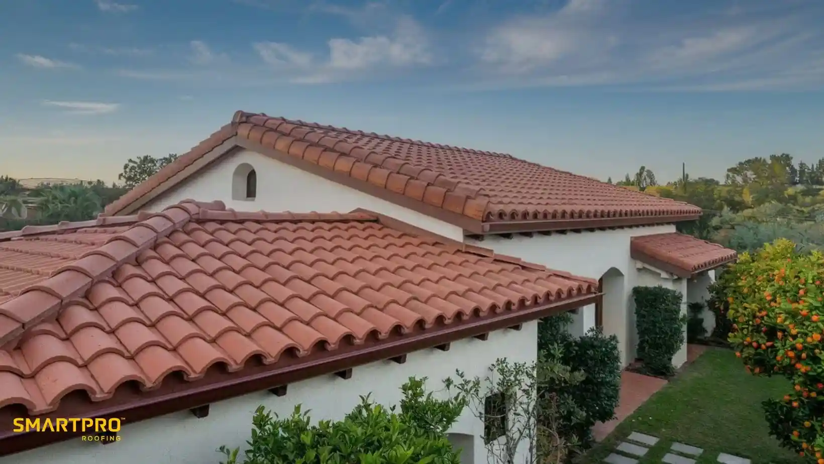 Spanish-style home with terracotta roof tiles, surrounded by lush greenery and an orange tree, set against a clear blue sky at dusk.