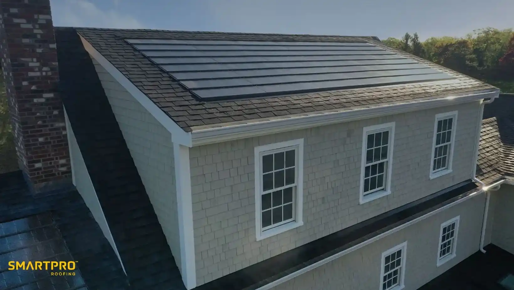 A house with solar panels integrated into a dark roof. The exterior is light-colored with multiple windows. The sky is clear, indicating sunny weather.