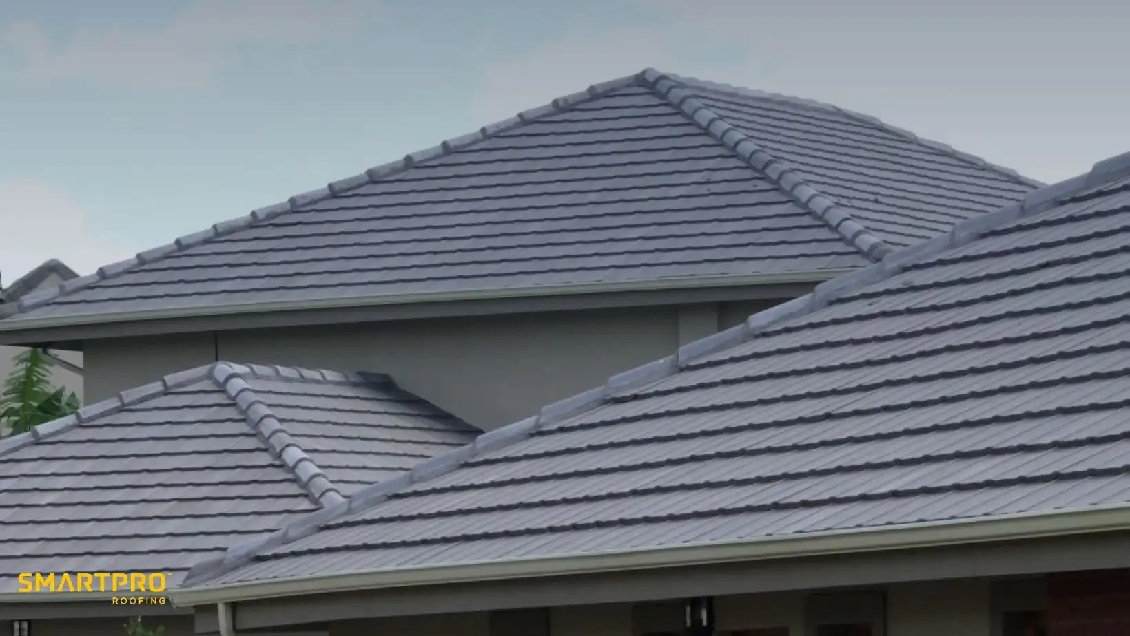 Steep, grey-tiled roofs overlap on a modern house against a clear sky. The structure conveys durability and efficiency. Logo at bottom left.
