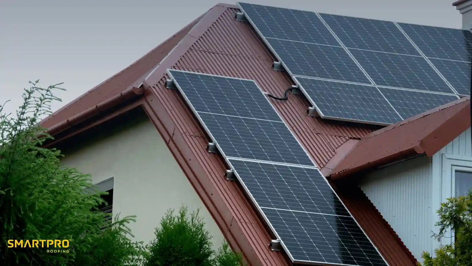 Rooftop with multiple solar panels on a red metal roof, surrounded by green trees, under a cloudy sky, conveying sustainability and modernity.