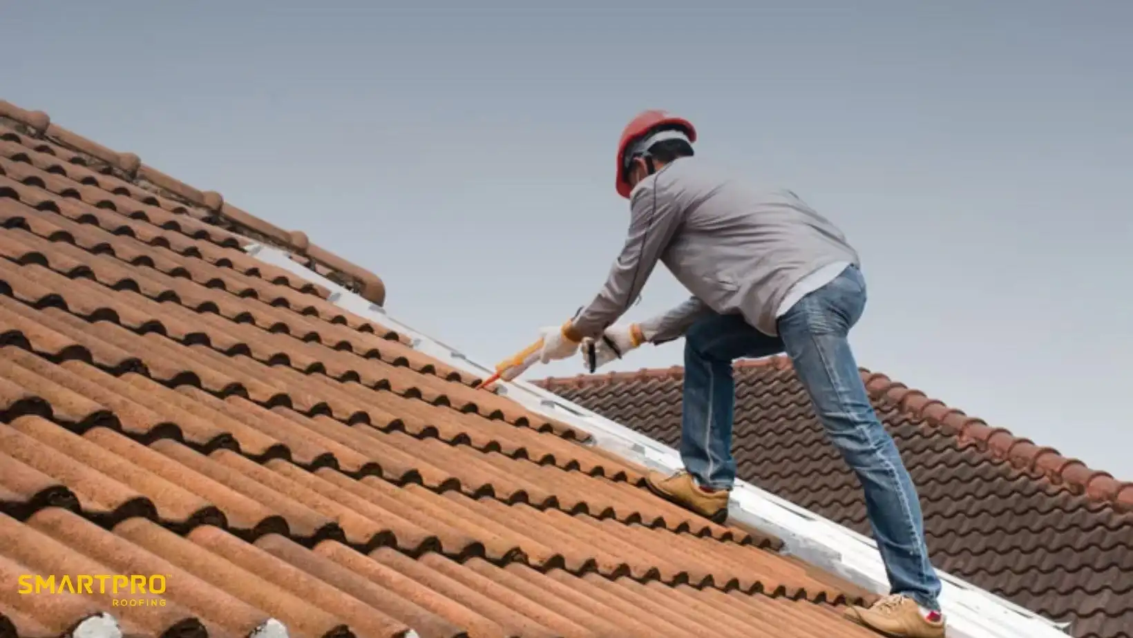 A worker in a red helmet repairs a tile roof under a clear sky, using a tool with careful precision. The scene conveys diligence and safety.