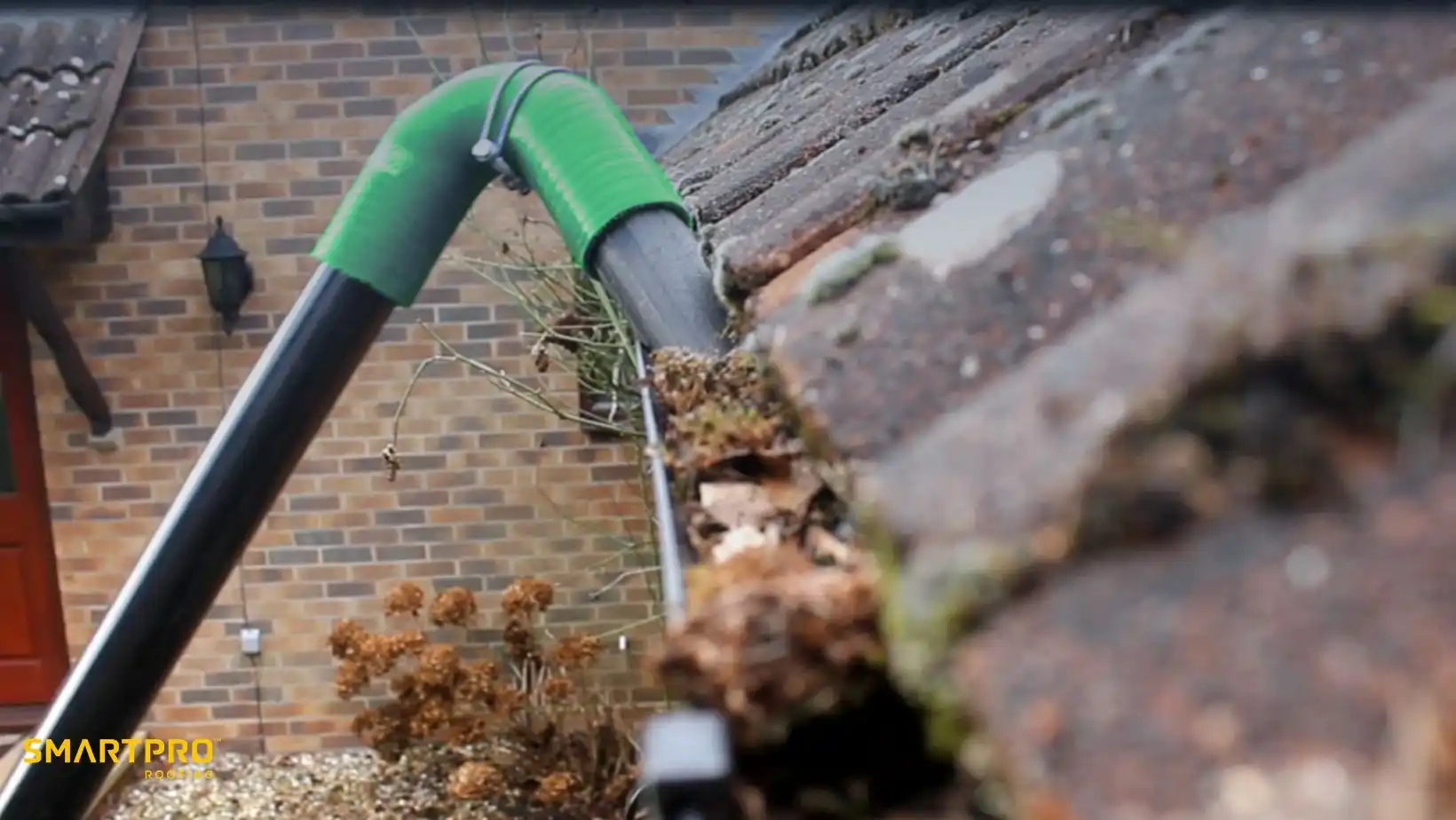 Close-up of a gutter being cleaned with a green and black tool. Debris and leaves are visible in the gutter, with a brick house in the background.