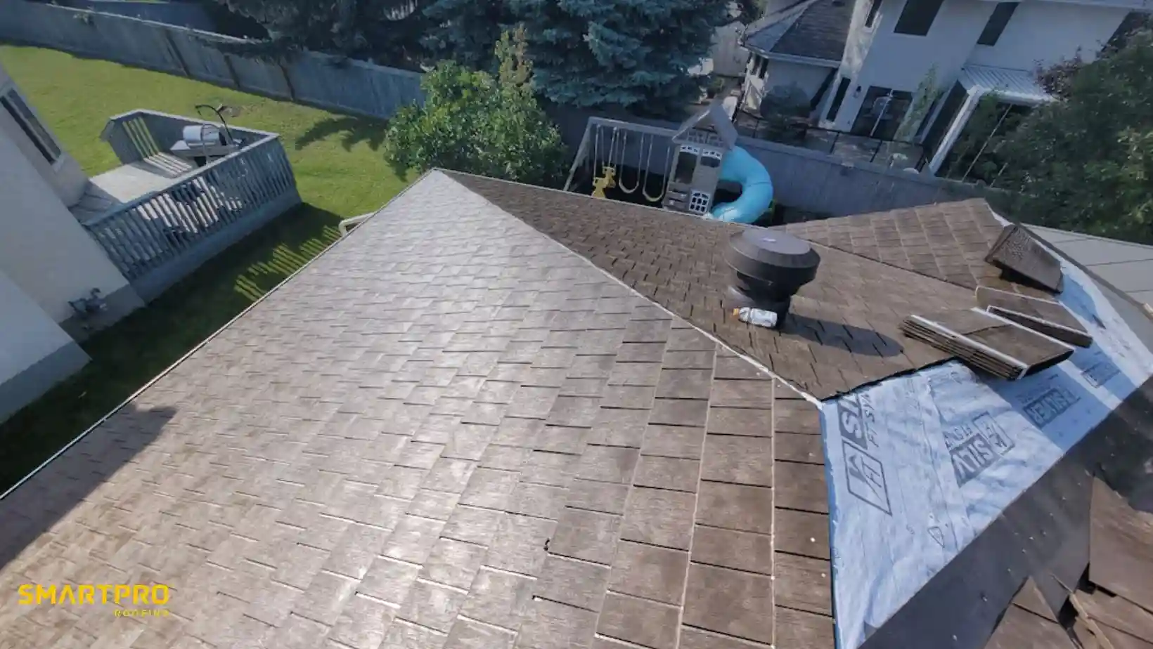 Aerial view of a house roof under repair, with brown shingles and blue underlayment visible. Backyard features a deck and children's playset. Bright, sunny day.