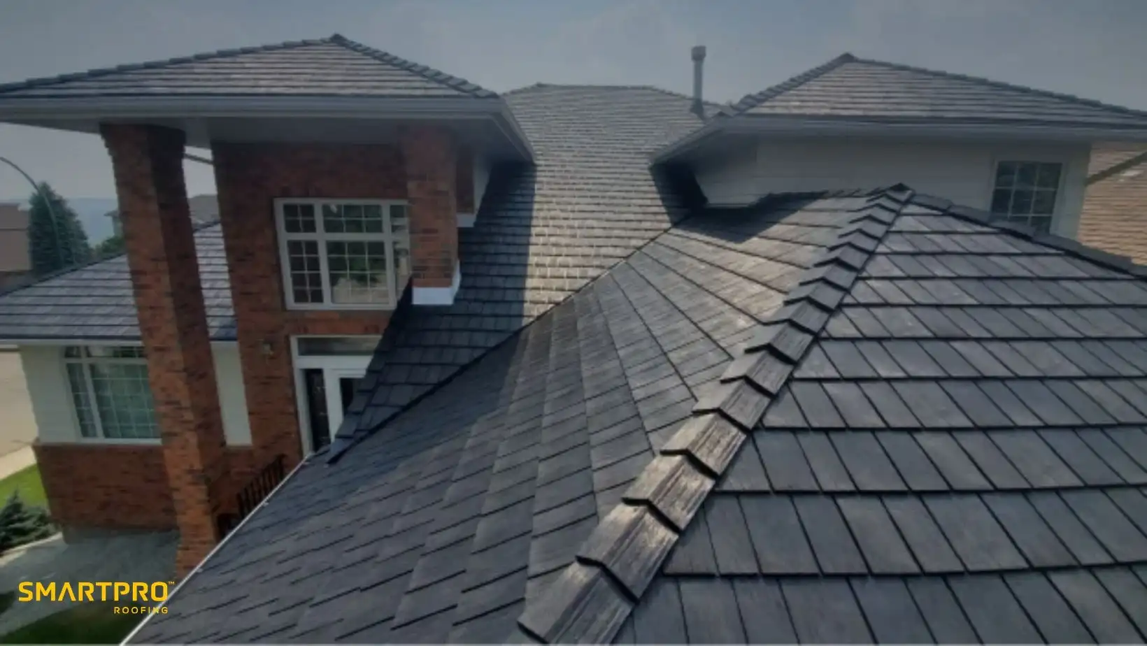 Aerial view of a house with dark gray, shingle-style roofing. The building has red brick walls and large windows, conveying a modern, sturdy appearance.