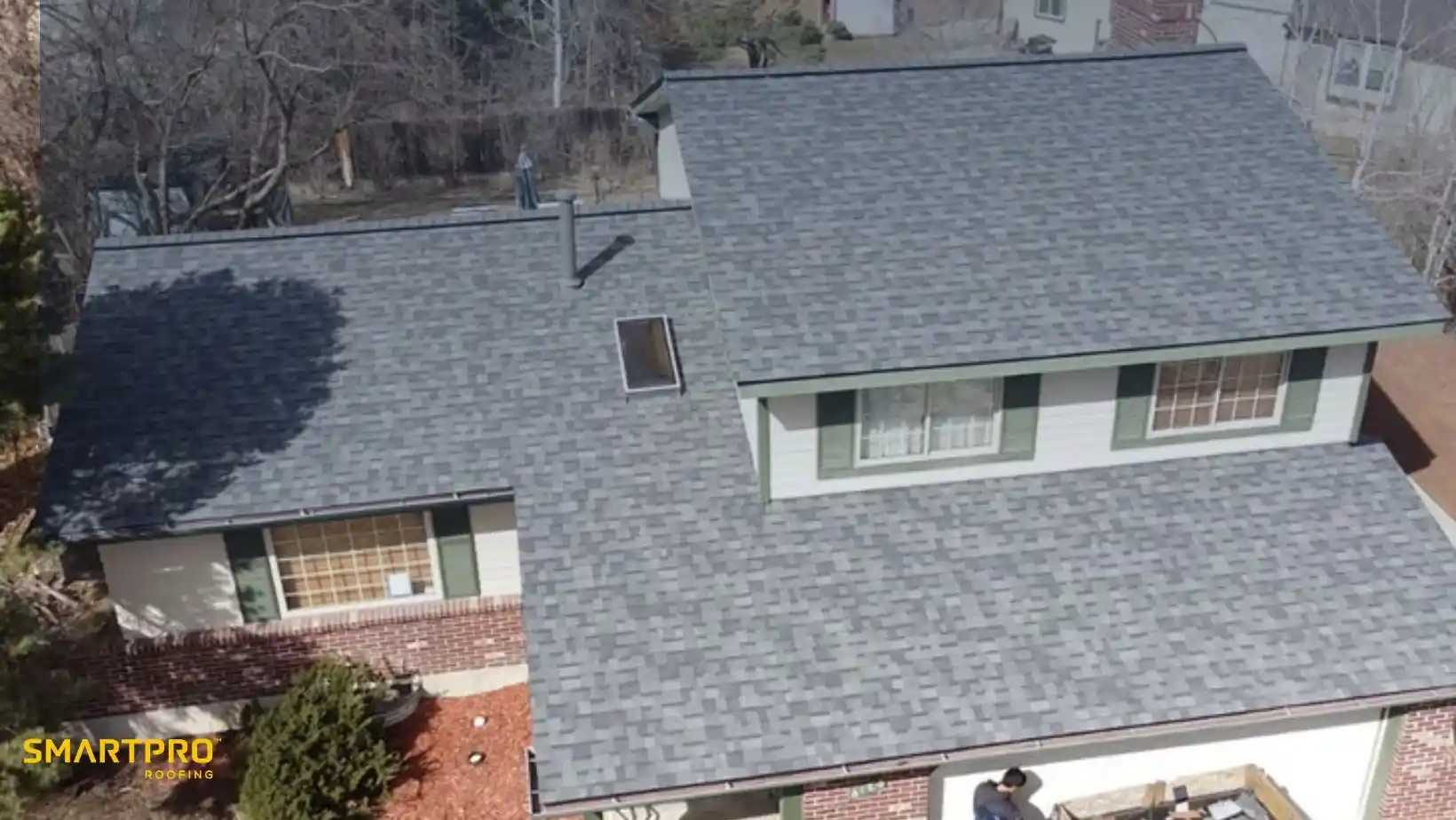 Aerial view of a house with a new gray shingle roof, green-trimmed windows, and red brick walls surrounded by trees, conveying a fresh, neat appearance.