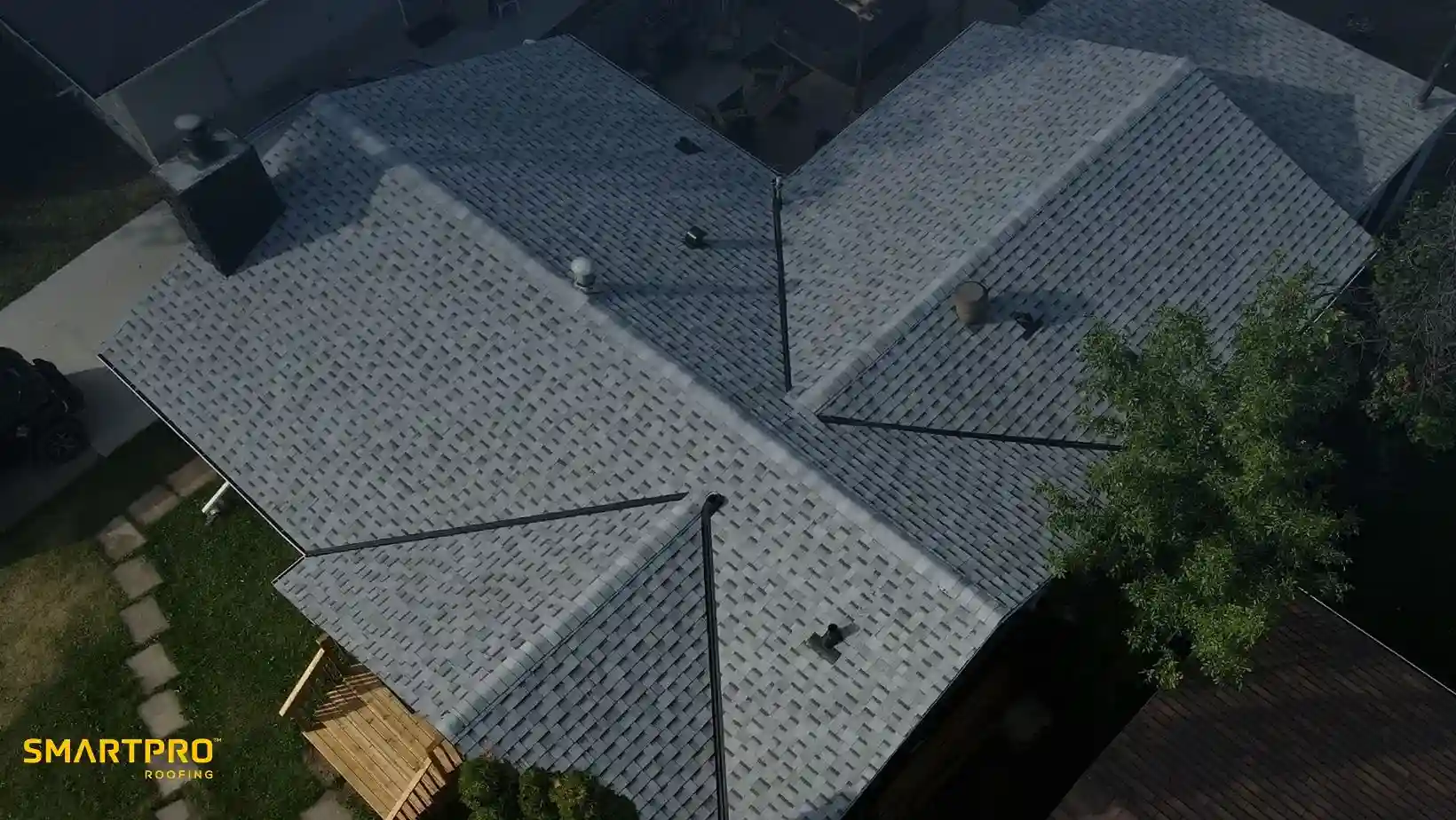 Aerial view of a house's roofing with a newly installed, light-colored shingle pattern, surrounded by trees and a driveway. The tone is professional.