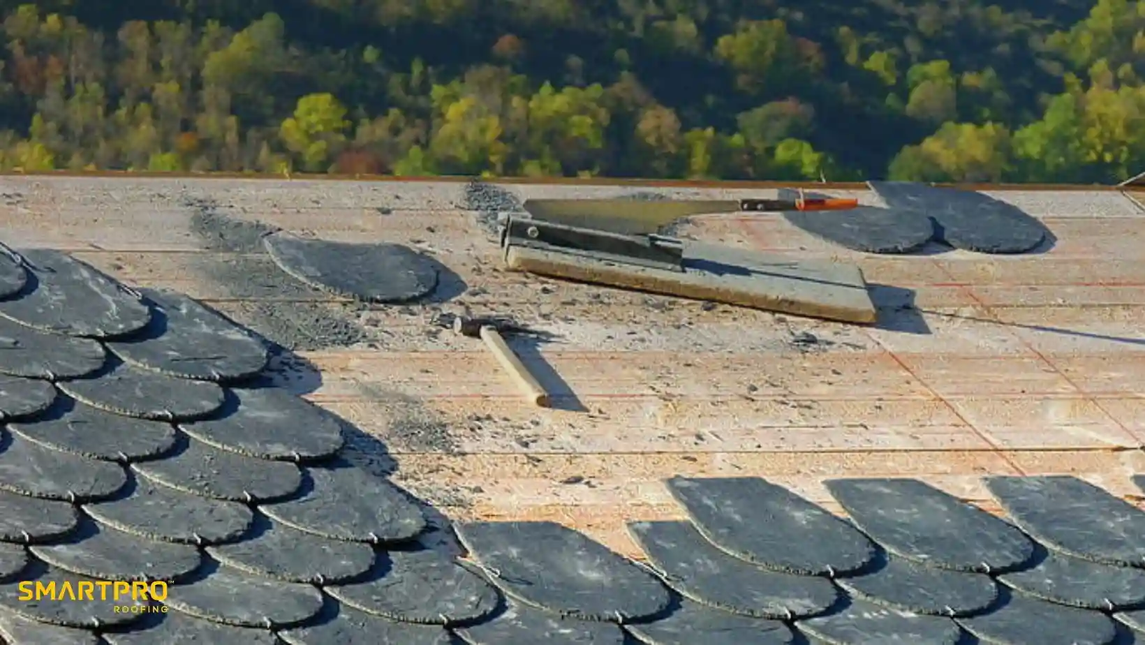 Close-up of a rooftop under construction, featuring gray scalloped shingles partially installed. Tools rest on the roof, set against a backdrop of lush, green trees.