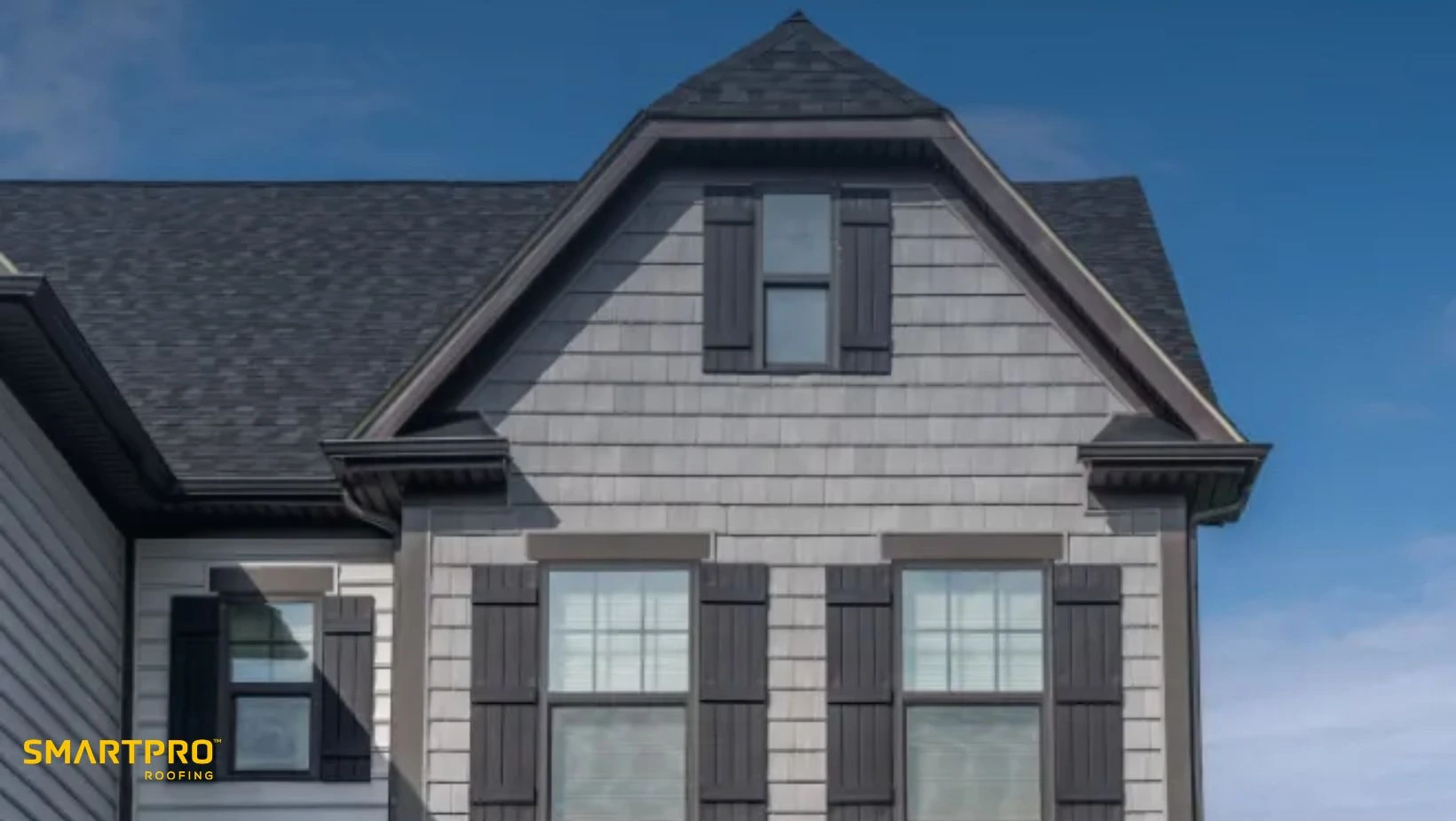 How Fiber Cement Compares to Composite and Wood Shingles 12 Gable roof house with gray siding and dark shutters under a clear blue sky. "SmartPro Roofing" logo is visible in the bottom left corner.